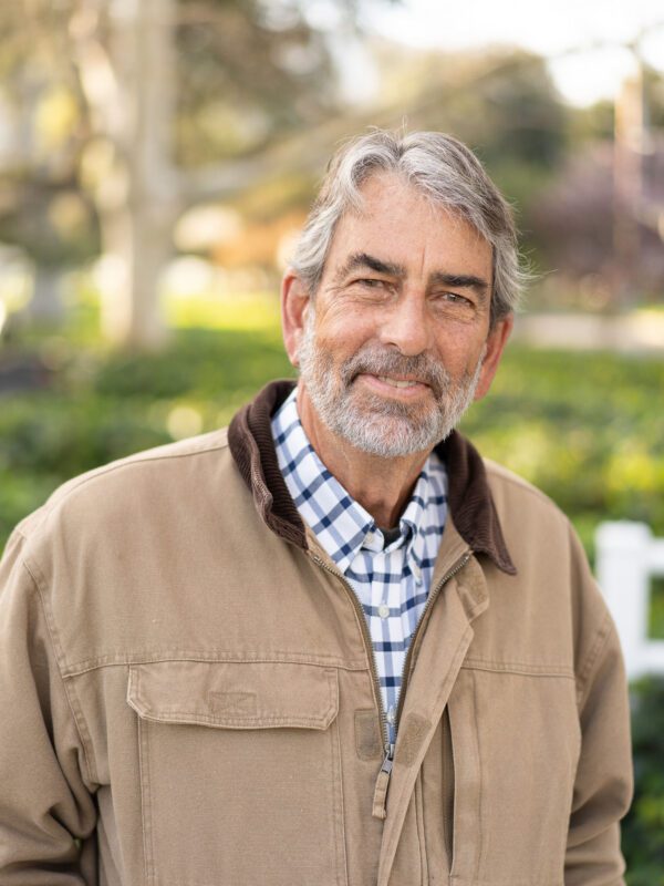 Outdoor photo of bearded older man smiling in coat with tree in background