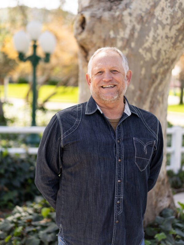 Outdoor photo of grey haired man with goatee smiling in dress shirt with tree in background