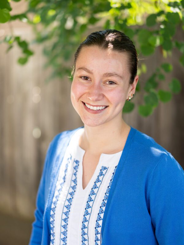 Outdoor photo of brunette woman smiling wearing a blue and white top with blurry leaves and fence in background