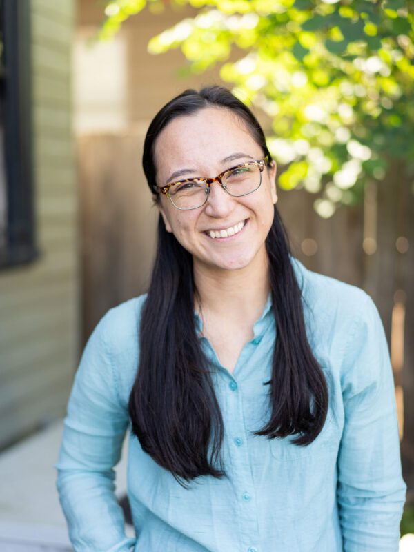 Outdoor photo of asian woman with glasses smiling with blurry leaves and fence in background