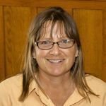 blonde woman with glasses wearing yellow shirt smiling in front of wooden door