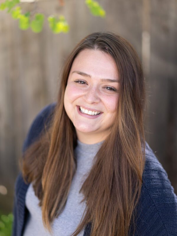 Outdoor photo of brunette woman smiling with blurry fense and leaves in background