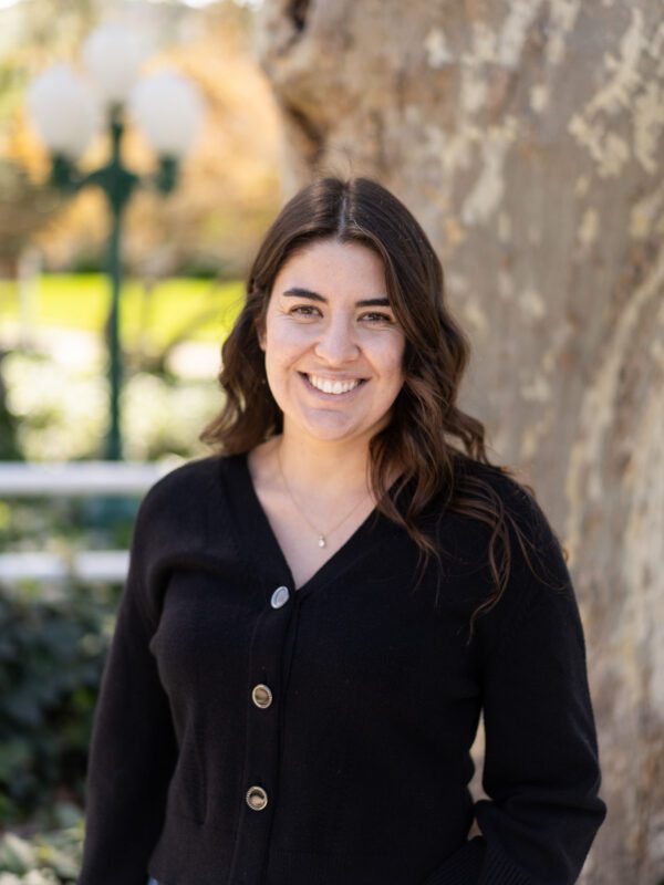 Outdoor photo of wavy haired brunette woman smiling with blurry tree in background